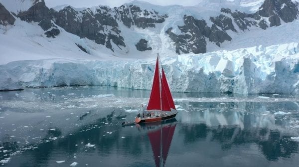 Sailboat with raised red sails moving toward glaciers and snowy mountains.