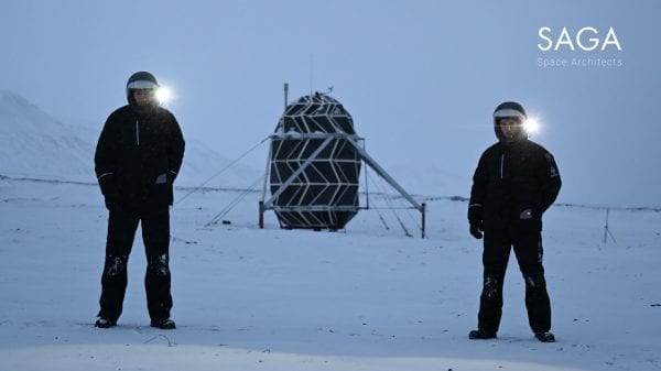 Sebastian and Karl wearing insulated suits and helmets standing in front of the Lunark module in Greenland, surrounded by snow.