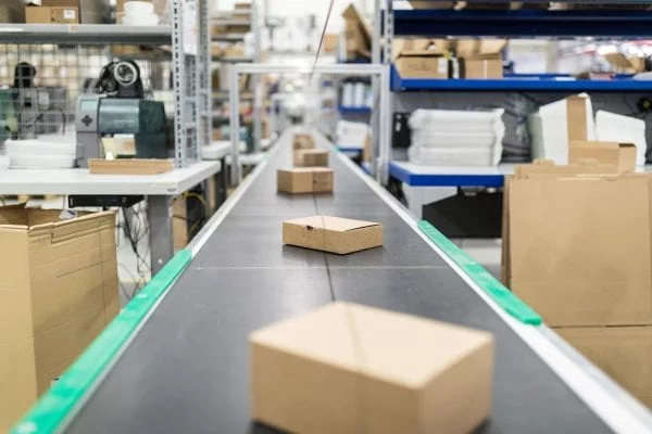 Brown boxes sliding along an assembly line through a factory between shelves stacked with supplies.