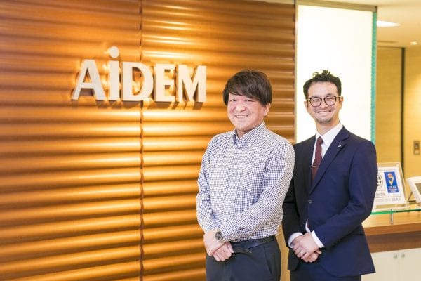 Two executives from AiDEM in front of company sign in an office