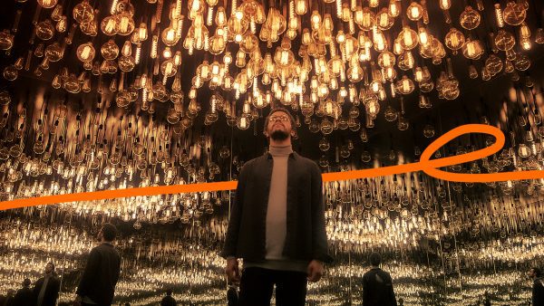 Lenovo Beyond Boundaries image - orange ribbon moving behind a man standing in a mirrored room with a ceiling covered in dozens of light bulbs
