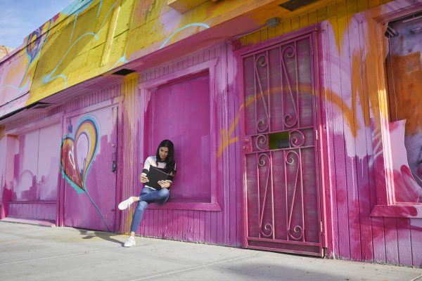 Woman sitting outdoors in front of walls and gates painted bright pink, using a Lenovo device