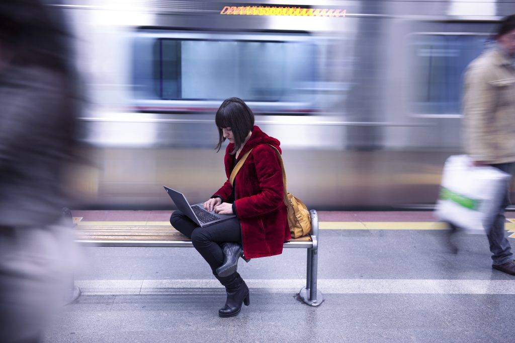 Brand image- Woman in a red coat sitting on a bench working on a laptop in a subway station.