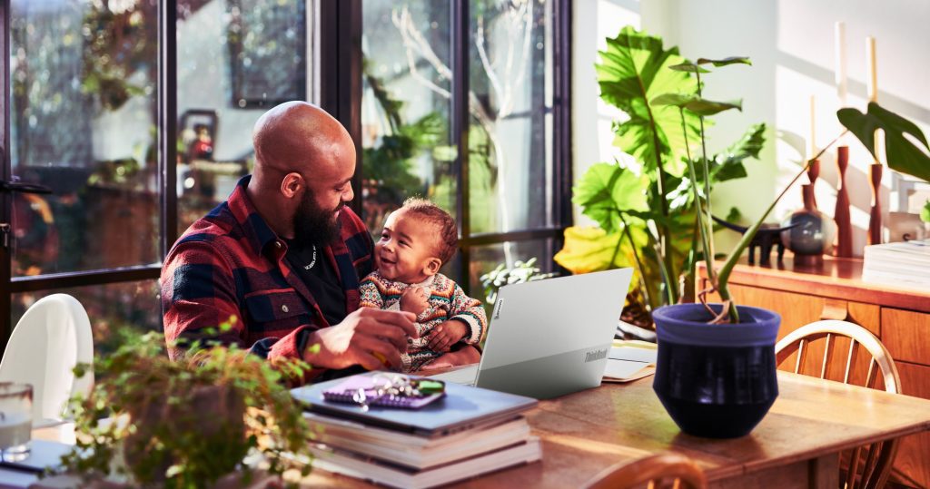 Father holding his baby, sitting at a table with a Lenovo laptop.