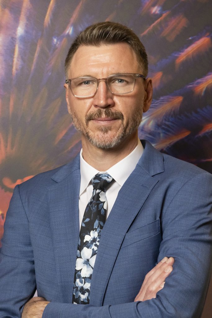 Portrait headshot of Matt Dobrodziej in a blue suit and tie, wearing glasses.