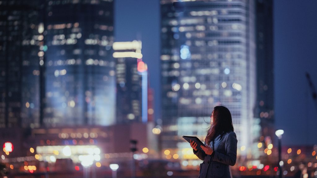 Brand image - Woman holding a tablet and looking at lit-up skyscrapers at night.