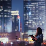 Brand image - Woman holding a tablet and looking at lit-up skyscrapers at night.